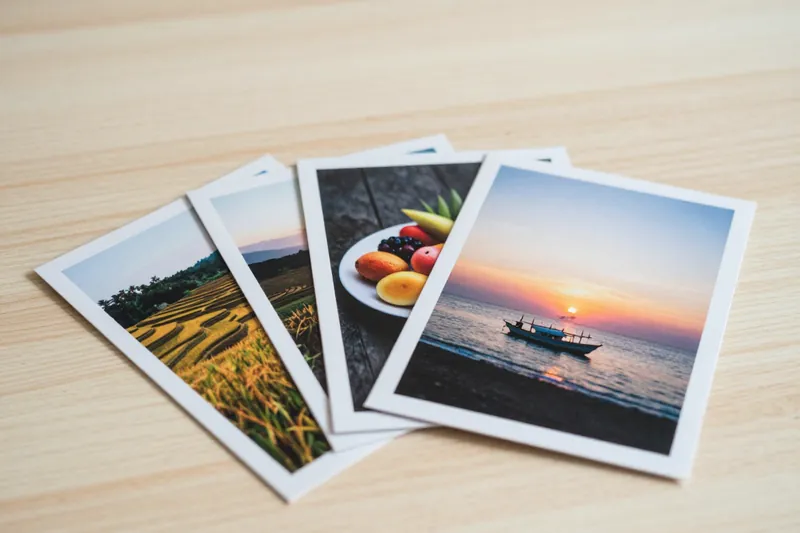 Cetakan foto berbingkai putih ukuran 3R, 4R, dan 5R di atas meja kayu: sawah terasering, sepiring buah, dan senja di pantai