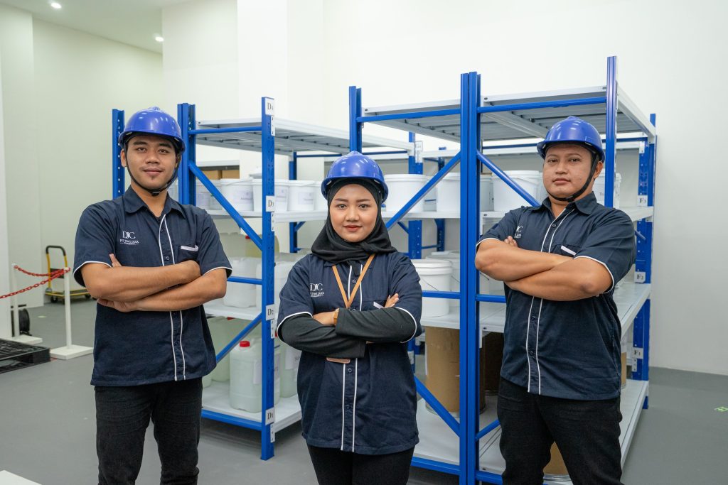 Three warehouse staff in hard hats in front of blue racks — Company Profile, PT Dwi Jaya Cosmedika
